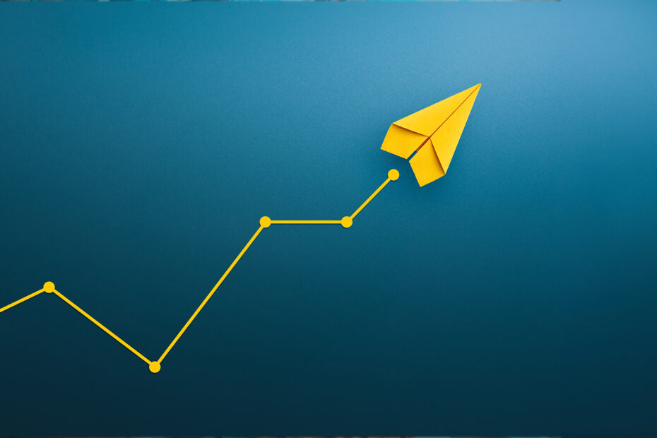 Paper airplane flying towards the sky in front of a blue wall.